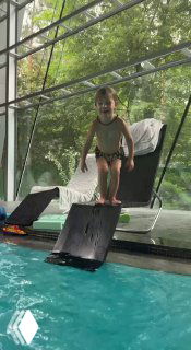 Ребёнок на бортике бассейна внутри комплекса на фоне больших окон с видом на лес, готовится к прыжку или игре в воде.