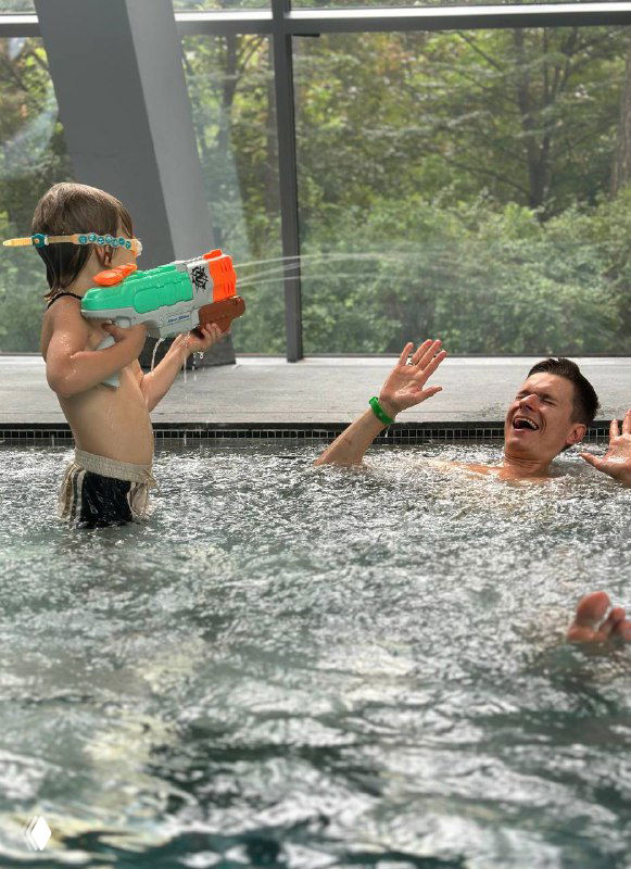 Мальчик с водяным пистолетом играет в тёплой воде рядом со взрослыми, снимок отражает семейный отдых и игровые моменты в джакузи/пруду.