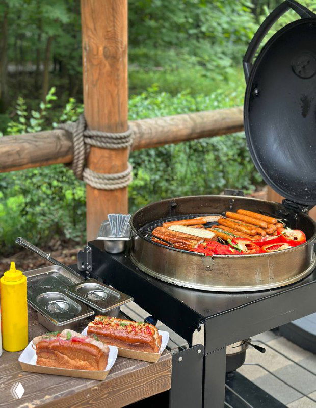 Уличный гриль с хот-догами и приправами на фоне летней террасы у ресторана комплекса, кадр подчёркивает наличие кафе и уличной еды для гостей.