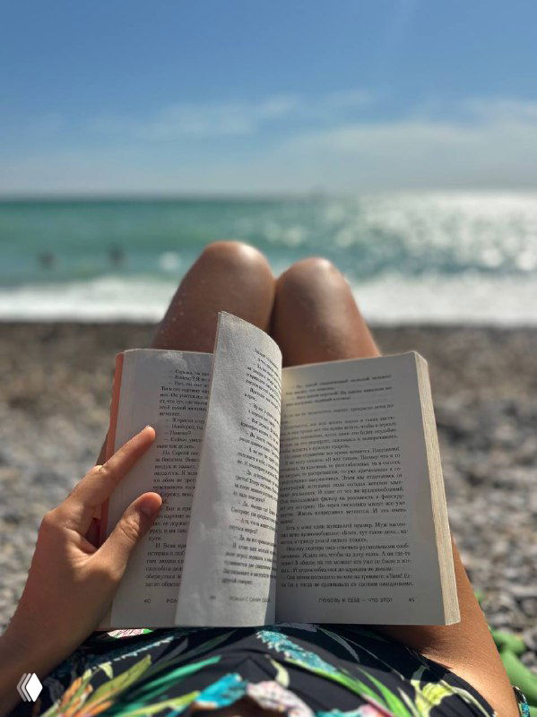 Крупный план раскрытой книги на коленях у человека на пляже, галька под ногами и море на заднем плане — спокойный момент чтения на отдыхе.