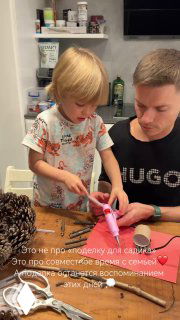 Ребёнок и отец за столом делают поделку; ребёнок использует клеевой пистолет, вокруг — материалы для творчества и шишки.