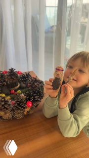 Ребёнок за столом держит готовую фигурку и улыбается; на столе — венок из шишек и украшения, плод семейного творчества.