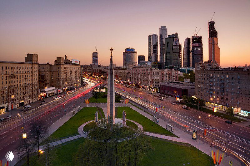 Вид на магистраль с монументом в центре кольца и небоскрёбы Москвы‑Сити на фоне заката, городской транспорт и развязки.