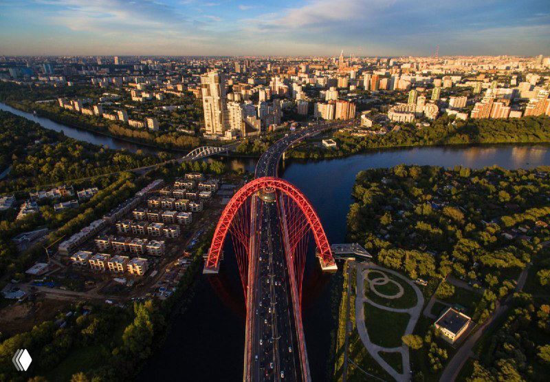 Аэрофото красного арочного моста через Москву‑реку, прилегающие парки и жилые кварталы, вид сверху на рекреационные зоны.