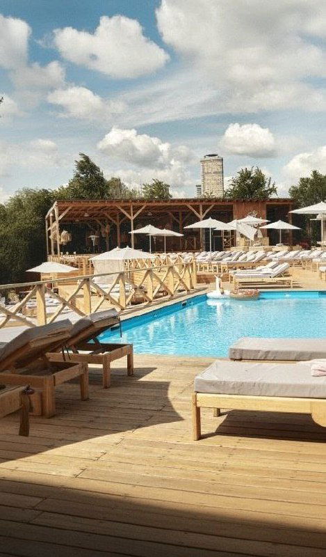 Пляжная зона с бунгало, деревянными лежаками и бассейном на настиле; непринуждённая летняя сцена для отдыха и мероприятий на открытом воздухе.