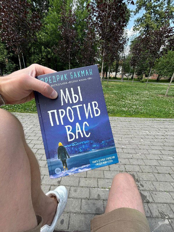 Рука держит книгу «Мы против вас» Фредерика Бакмана на фоне летнего парка и скамейки, видны ноги и городская зелень.
