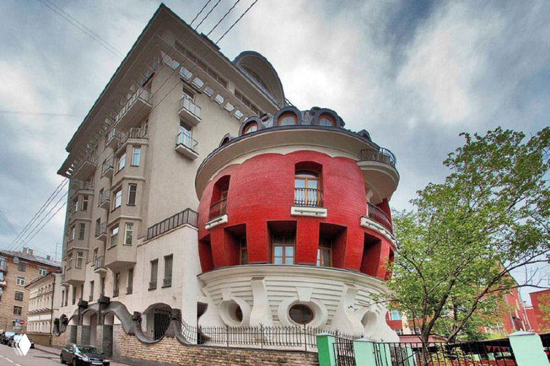 Другой ракурс красного дома‑яйца на Машковой в окружении жилых зданий и деревьев; видно цилиндрическое тело, керамическую отделку и входы в основание