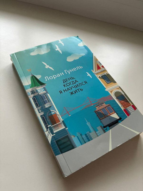 Книга Лорана Гунеля «День, когда я научился жизни» лежит на подоконнике; обложка с яркой иллюстрацией городского пейзажа и небом.