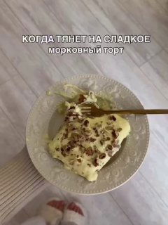 Кусок морковного торта с белым кремом и грецкими орехами на белой тарелке, вид сверху на светлом деревянном полу.