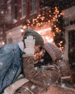 Молодая пара в зимней одежде обнимается и целуется на заснеженной улице вечером; вокруг падает снег и горят праздничные огни на фоне.