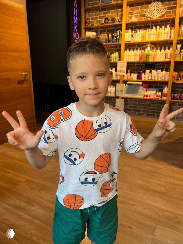 Фронтальный портрет мальчика после стрижки; улыбается и показывает жесты, видно стильную футболку и интерьер салона на фоне.