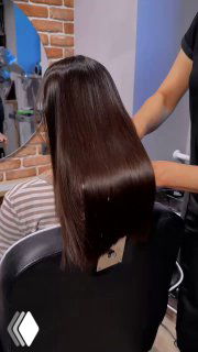 Клиент в салоне с результатом кератинового выпрямления: длинные тёмные волосы с ровной глянцевой поверхностью, мастер держит прядь.