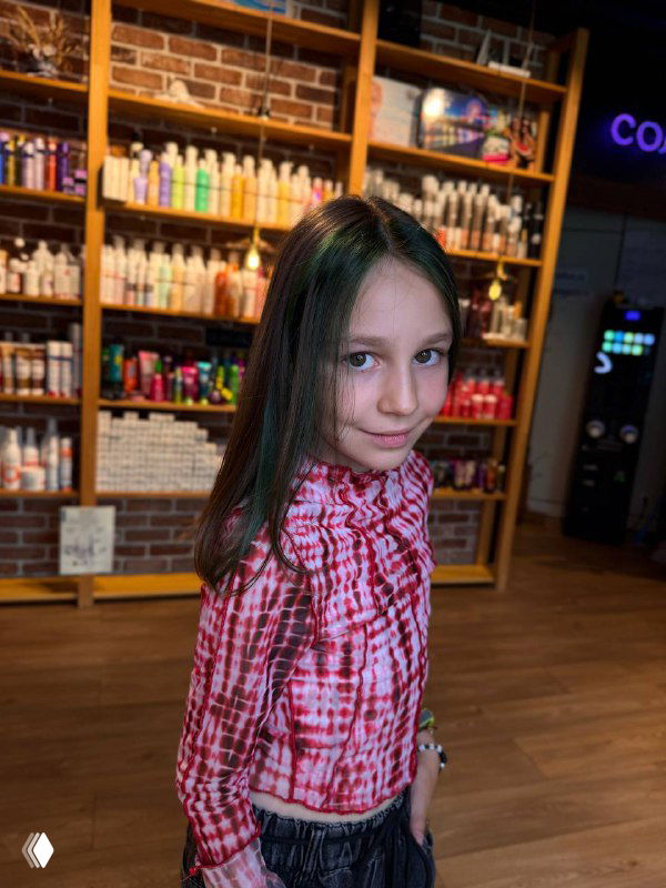 Фотопрофиль девочки с зелёными кончиками волос и модной блузкой; на заднем фоне, полки с товарами салона.