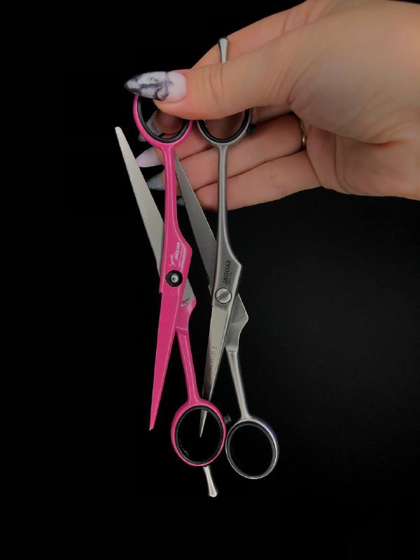 Две парикмахерские ножницы — розовая и серебристая — в женской руке на тёмном фоне; демонстрация инструментов для стрижки и укладки.