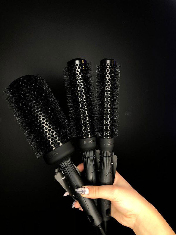 Набор трёх круглых щёток для сушки и укладки волос в руке на чёрном фоне; изображены рабочие аксессуары для причёсок и салонов.
