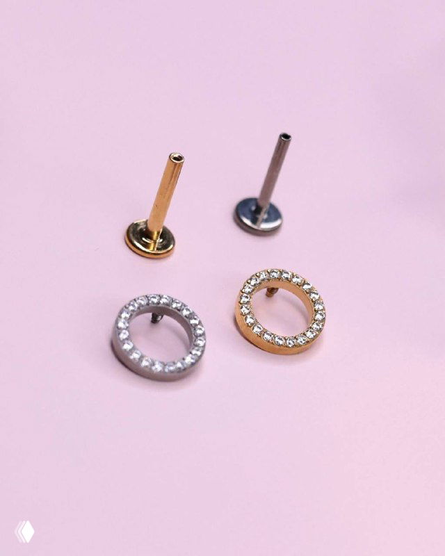 Круглые титановые накрутки с инкрустацией кристаллов и соответствующие стержни push-in на пастельном фоне, видна зеркальная поверхность металла.