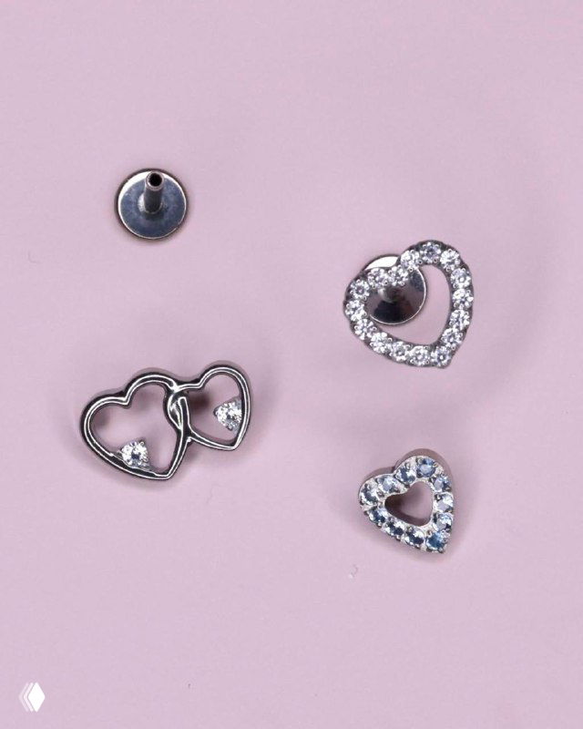 Несколько титановых накруток в форме открытых и инкрустированных сердец рядом со стержнем push-in на мягком розовом фоне, подчёркнут блеск кристаллов.