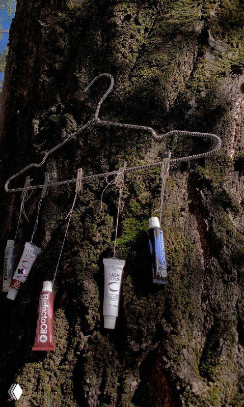 Фотография: несколько тюбиков профессиональной краски для ресниц и бровей, подвешенные на металлической вешалке на фоне коры дерева — товарная демонстрация.