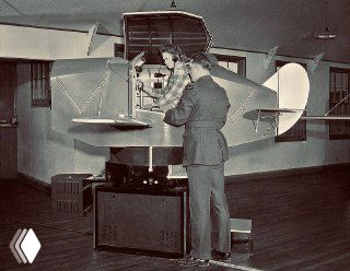 Ранний Link Trainer на подвижной платформе: вид сбоку с открытой кабиной и инструктором рядом, историческая чёрно‑белая фотография учений.