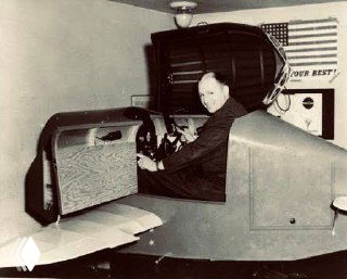 Исторический кадр с Link Trainer: мужчина в кабине тренажёра демонстрирует органы управления, на заднем плане элементы учебного зала и флаг.