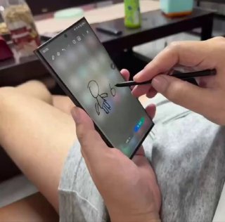 Рука с стилусом рисует простую схематичную фигуру на экране смартфона, видны ноги в шортах и фон с предметами на столе