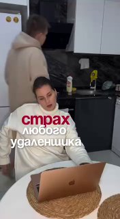 Кадр домашнего офиса: женщина сидит за ноутбуком за кухонным столом, на заднем плане человек у кухонного гарнитура; бытовая обстановка.