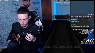 Стример держит гитарный контроллер перед монитором с интерфейсом Guitar Hero; видны дорожки нот, индикаторы и чат.