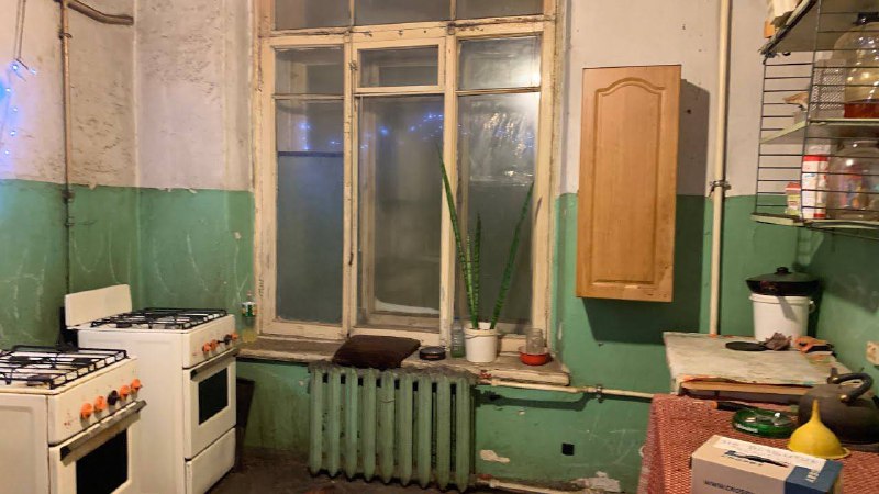 Старая кухня с газовой плитой, раковиной, радиатором и высветленным окном — реальный бытовой фон, создающий непринуждённую атмосферу.