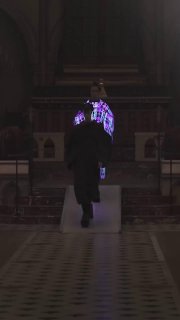 Кадр с показа в тёмном зале: один из образов подсвечен фиолетовыми LED‑элементами на спине плаща, модель идёт по проходу между рядами зрителей.