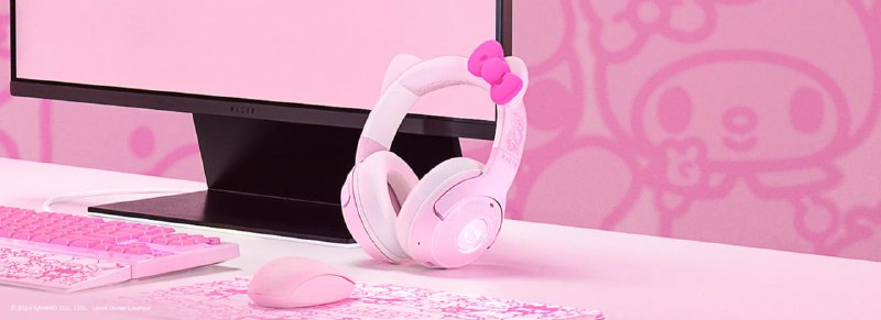 Розовые накладные наушники Hello Kitty на рабочем столе рядом с монитором, часть комплекта коллаборации Razer в фирменном дизайне.