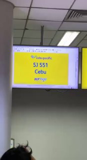Жёлтый информационный экран в аэропорту, отображающий номер рейса и направление «Cebu», видна часть потолка и колонна терминала на фоне.
