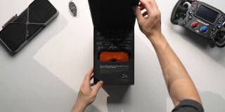 Руки открывают коробку с аксессуарами Bigscreen Beyond 2 на светлом столе; виден оранжевый чехол и часть упаковки рядом с контроллером.