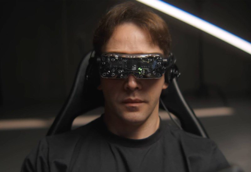 Крупный план мужчины в темной одежде, он надевает или уже носит VR-гарнитуру Bigscreen Beyond 2; видны лицевые сенсоры и фронтальная панель.