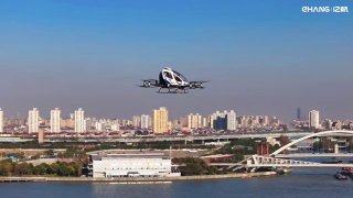 Беспилотное летающее такси над городской набережной и мостом, на фоне виден городской горизонт, река и здания под ясным небом.