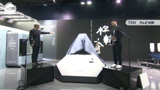 Сцена выставки с накрытым прототипом робо‑коня на подиуме, двое людей по бокам и задний экран с надписями
