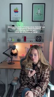 Девушка сидит за столом в домашнем офисе и держит телефон; на заднем плане полка с рамками, микрофоном, лампой и подсветкой.