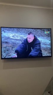 Телевизионный экран на стене с крупным кадром мужчины в куртке, сидящего в полевой местности; кадр иллюстрирует сцену телепередачи.