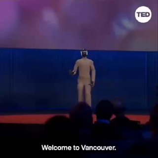 Робот‑гуманоид стоит на сцене TED со спины; видны логотип TED в углу, тёмная аудитория и надпись «Welcome to Vancouver» на экране.