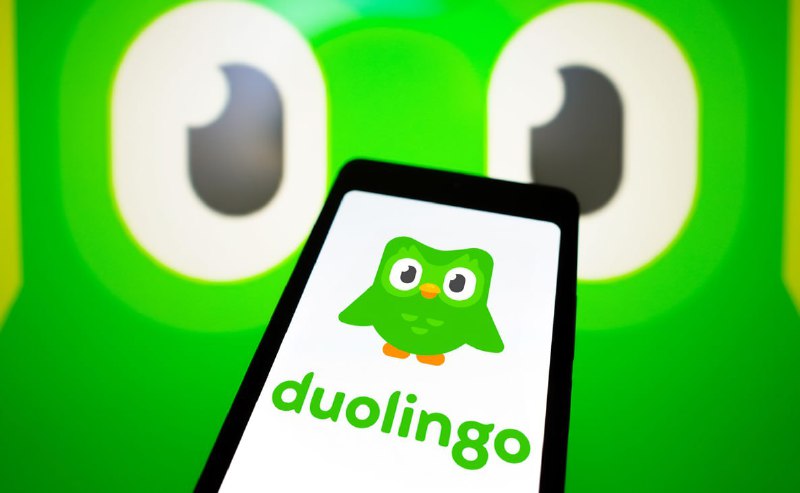 Логотип Duolingo на экране смартфона на фоне большого зелёного изображения совы; визуализация новости об автоматизации и ИИ.