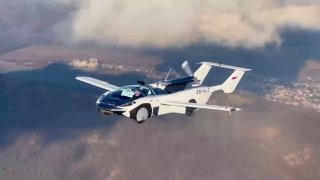 Фото: белый летающий автомобиль в полёте над местностью, видны выдвижные крылья, фюзеляж и кабина — демонстрация модели.