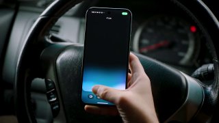 Рука держит смартфон в салоне автомобиля, кадр с мобильным интерфейсом на фоне руля и приборной панели.