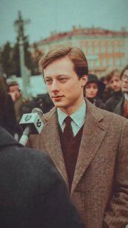 Молодой мужчина в пальто отвечает в микрофон на городской площади: кадр уличного интервью с прохожими и зданиями на заднем плане.