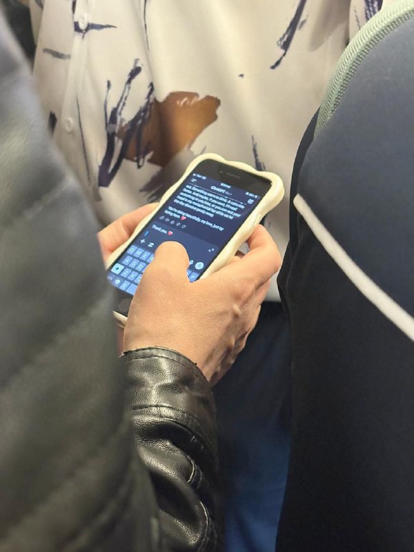 Рука пассажира в вагоне метро держит смартфон с открытым чатом; на экране видно текст диалога с чат‑ботом, вокруг фрагменты других пассажиров и салона.