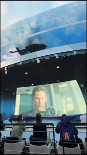 Изогнутый панорамный экран в зале, на котором проигрывается фрагмент фильма, видна вертолётная тень и зрители в переднем ряду в кадре.