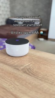 Круглое бело‑чёрное голосовое устройство на деревянном столе в интерьере; размытый фон и текст об ИИ и учителях.