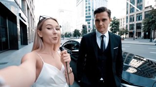 Женщина с микрофоном делает селфи-интервью рядом с деловым мужчиной в костюме и дорогой машине на городской улице.