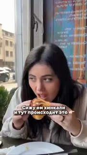 Кадр из видео: человек в кафе за столом у окна ест хинкали руками над тарелкой; на заднем плане видна улица и вывески.