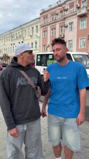Двое взрослых мужчин стоят на городской улице в повседневной одежде, беседуют на фоне исторических домов и автобуса.