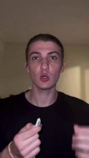 Вертикальный кадр видео: молодой человек в комнате обращается к камере и держит маленький предмет на фоне нейтральной стены.