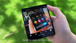 Руки держат компактный квадратный смартфон с видимым интерфейсом приложений на экране; устройство на размытом зелёном фоне, снято крупным планом.